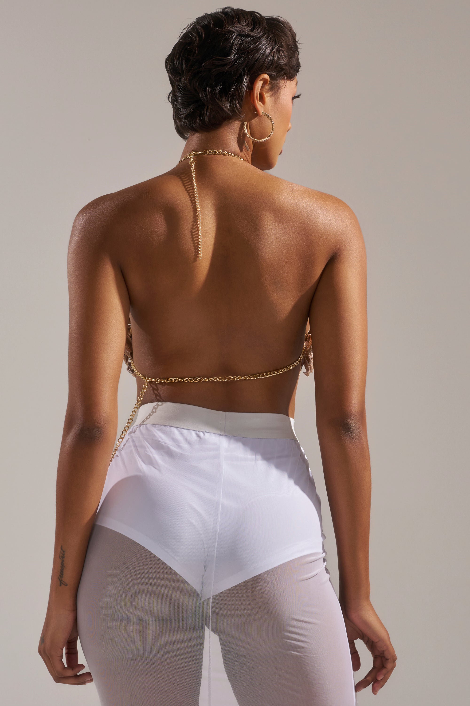 A woman with short hair and hoop earrings stands against a neutral background, wearing the ROSES BRALETTE—a gold chain bralette—paired with white sheer high-waisted pants, her bare back visible as her hand rests at her side. 5