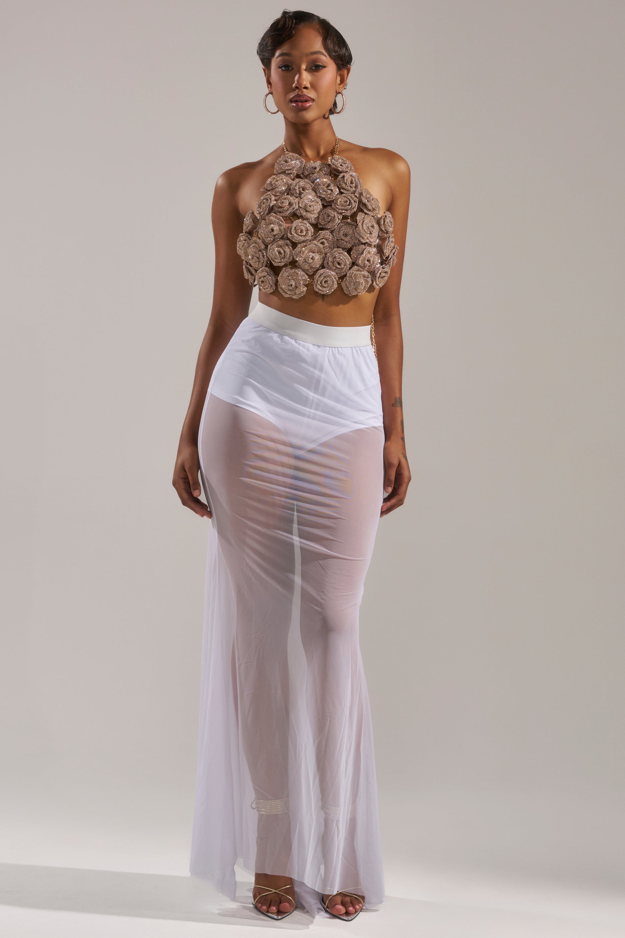 A woman with short hair and hoop earrings poses against a plain background wearing the ROSES BRALETTE—a halter neck bralette with large, textured floral designs in a neutral tone—paired with a sheer white floor-length skirt. 7