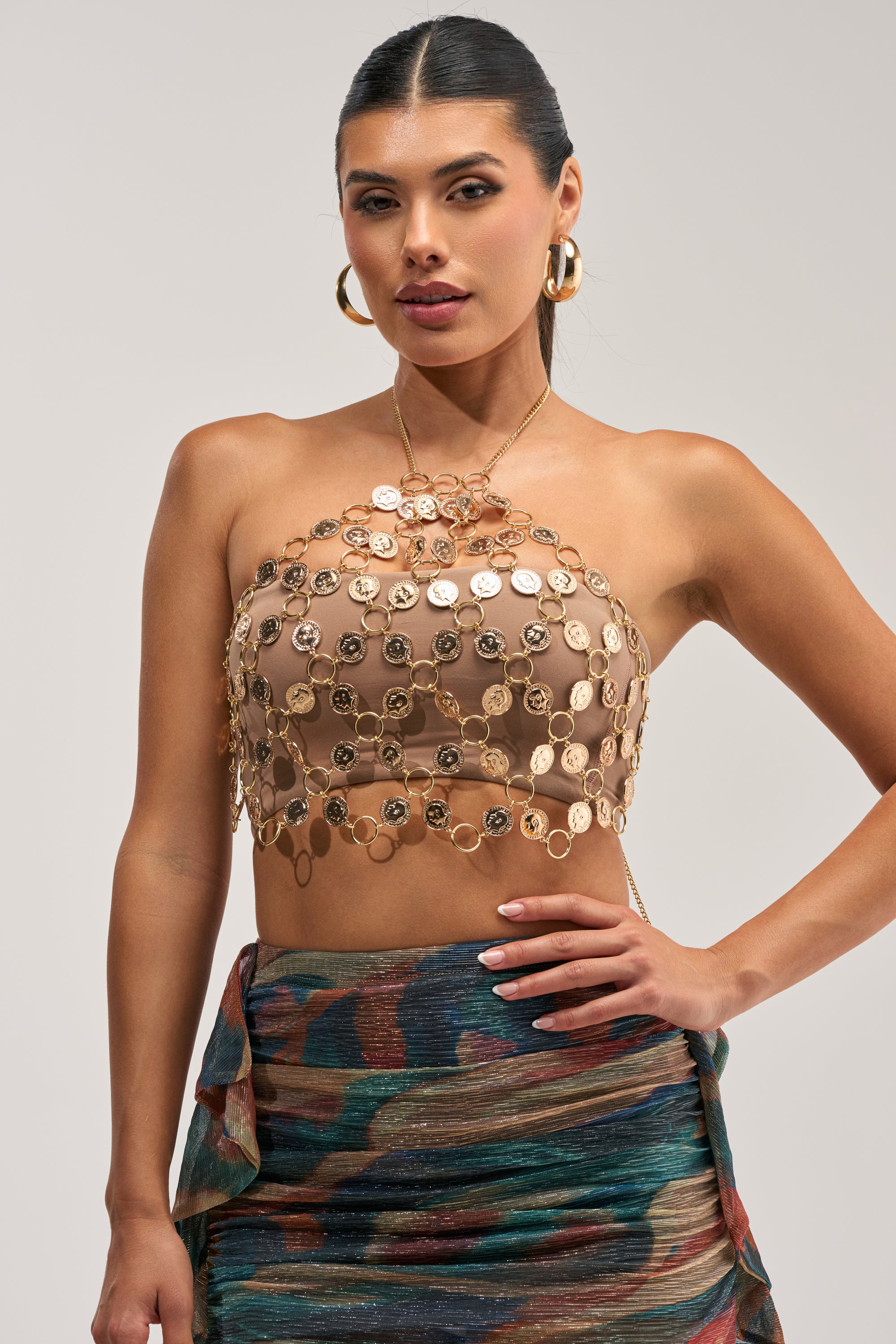 A woman poses against a plain background, wearing a tan tube top layered with the COUNT THAT COIN BODY CHAIN—a decorative gold coin-link body chain—paired with gold hoop earrings and a multi-colored metallic patterned skirt featuring a ruched, wrap-style design.