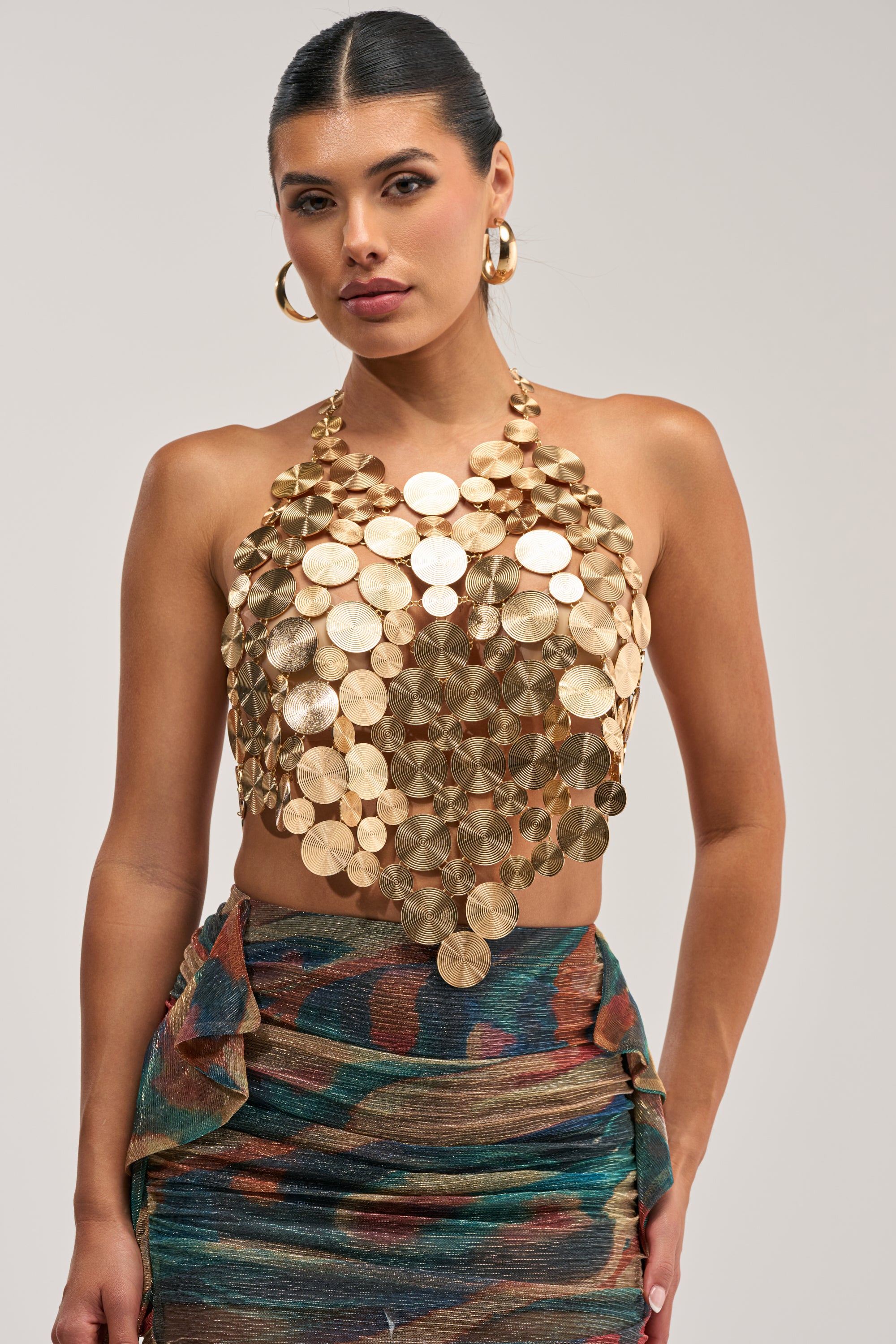 A woman with dark hair in a slicked-back bun poses against a white background, wearing the GOING IN CIRCLES METAL CHAIN TOP—a halter style constructed from layered, textured gold-toned metal discs—paired with a colorful, metallic ruched midi skirt and large gold hoop earrings.