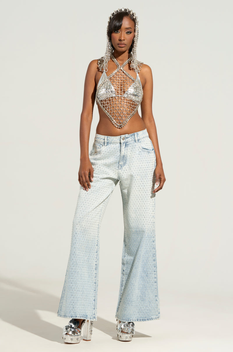 A model showcases a silver chainmail halter top paired with the IYKYK Luxe Body Chain glinting beneath, complemented by a matching headdress and light blue wide-leg jeans featuring metallic accents. She confidently stands on silver platform heels against a plain background, her outfit shimmering with bling.