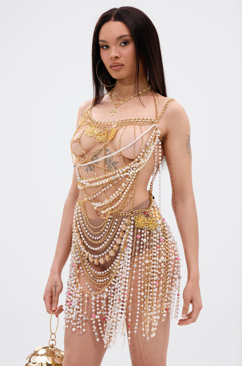 A woman with long dark hair wears the NAMIKA LAYERED CHAIN AND PEARL MINI DRESS, showcasing gold-tone chain links and draped pearls. She holds a gold bag, revealing tattoos on her chest and arms against a white background.