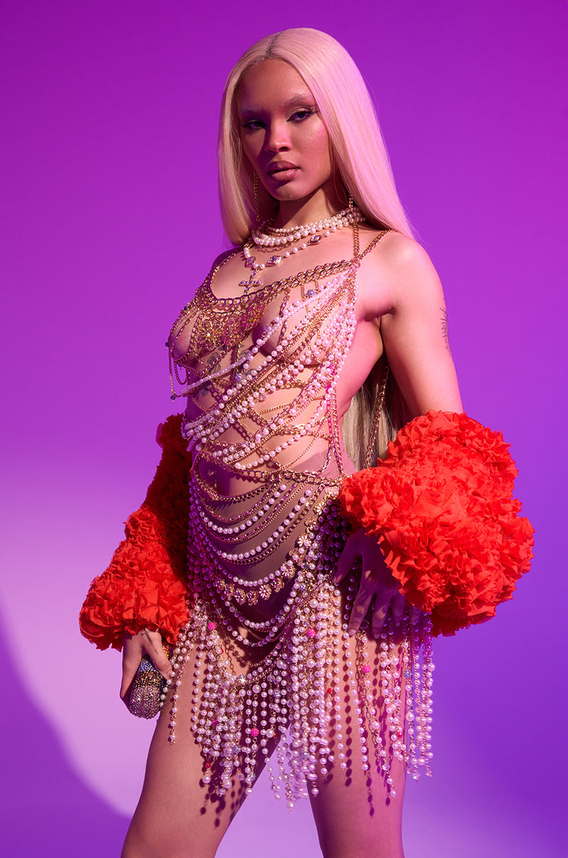 A woman with long blonde hair poses against a purple backdrop, wearing the NAMIKA LAYERED CHAIN AND PEARL MINI DRESS with bold red ruffled sleeves. She holds a sparkling clutch and gazes confidently at the camera.