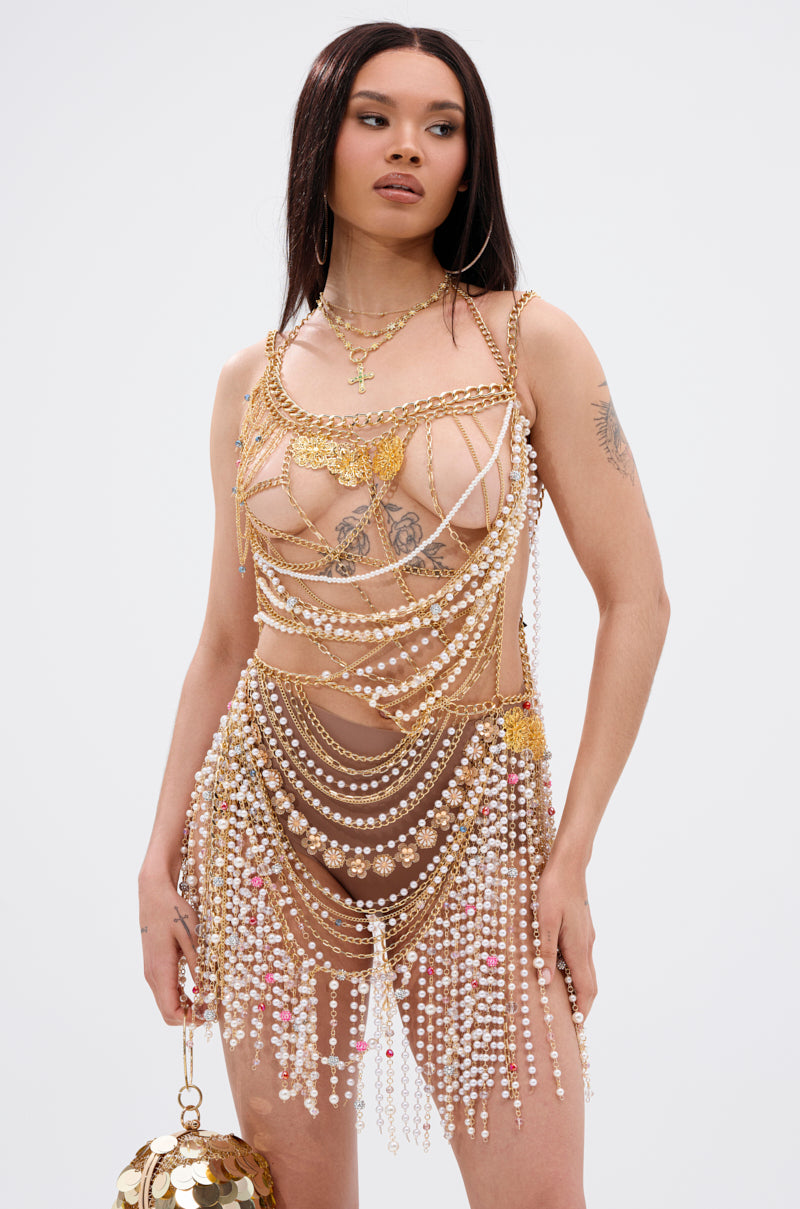 A woman exudes confidence in the NAMIKA LAYERED CHAIN AND PEARL MINI DRESS, a nude mesh mini dress with gold chains and pearl accents. She pairs it with a coin-detailed purse, sleek straight hair, and visible tattoos for a striking look.