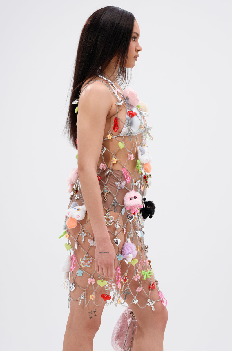 A woman with straight dark hair models the NOT SO TYPICAL CHAIN MINI DRESS WITH CHARMS, a sheer mesh mini dress embellished with playful charms, standing against a plain white background.