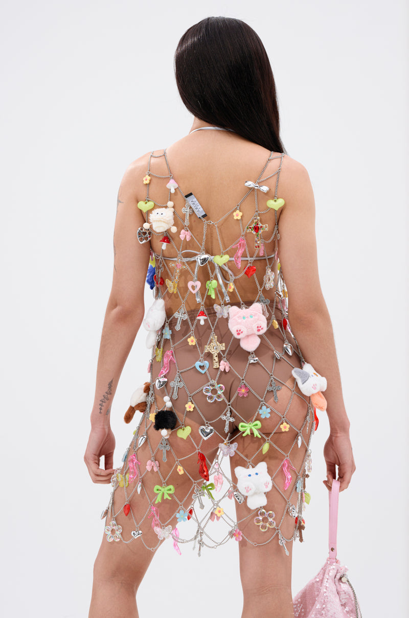 A person with long dark hair is shown from the back, wearing the NOT SO TYPICAL CHAIN MINI DRESS WITH CHARMS adorned with plush toys and bows. They hold a pink glittery handbag, standing against a plain white background.