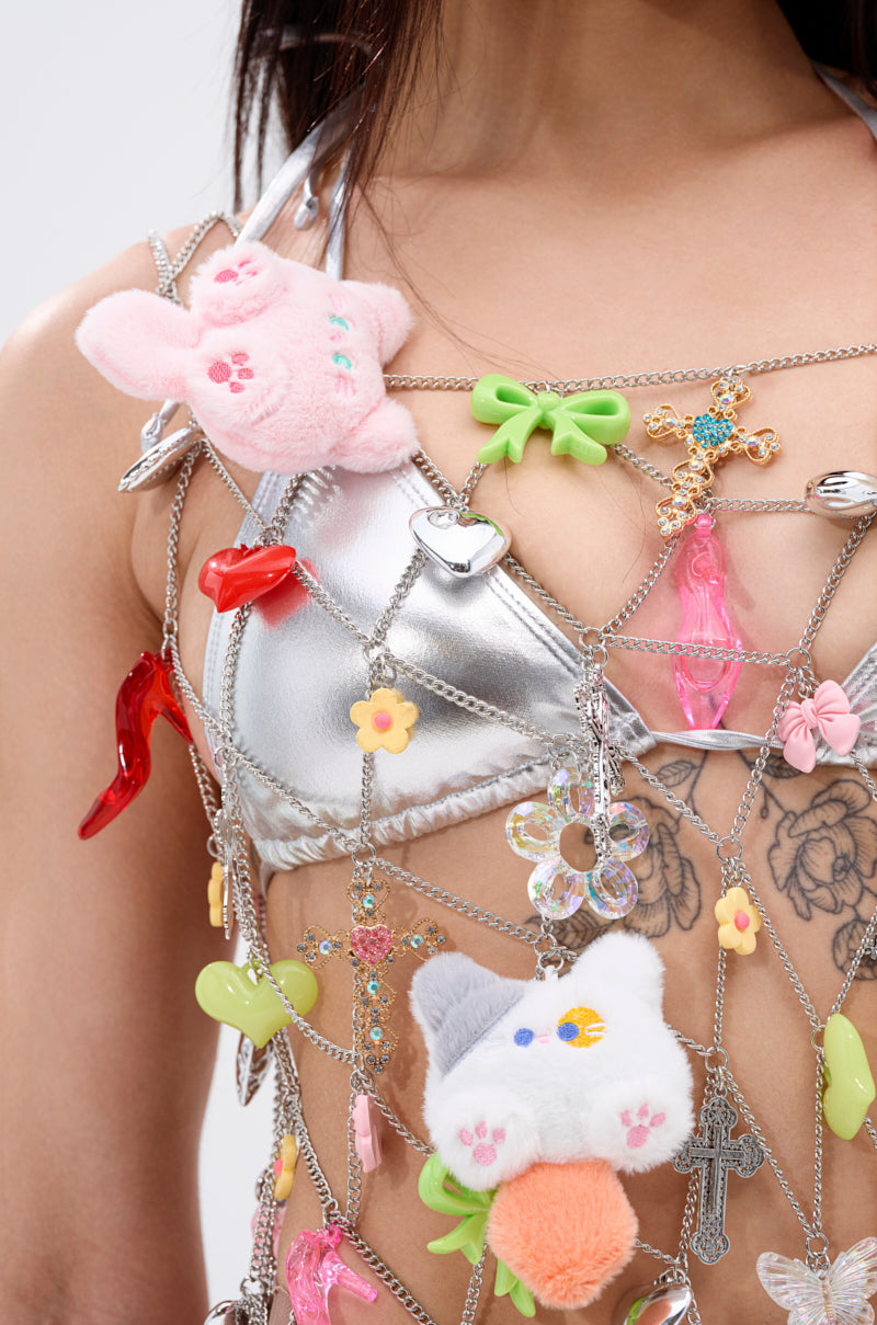 A person wearing the NOT SO TYPICAL CHAIN MINI DRESS WITH CHARMS, featuring eclectic charms, plush toys, and trinkets over a silver bikini top, with chest tattoos visible. The cropped image shows their torso and part of their face.