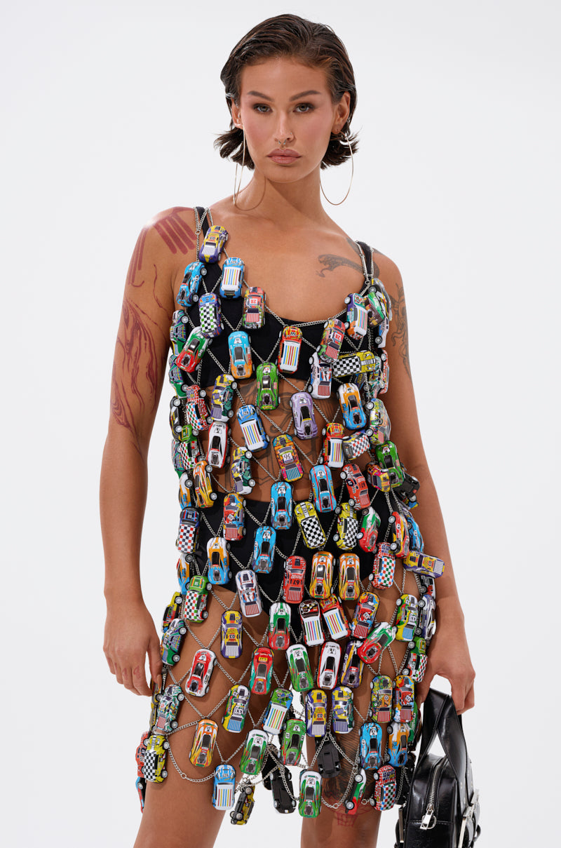 A woman with wet-look hair and hoop earrings models the CAN'T CATCH ME CAR CHAIN MINI DRESS, made of colorful toy cars on mesh. She stands confidently holding a black handbag against a neutral background.