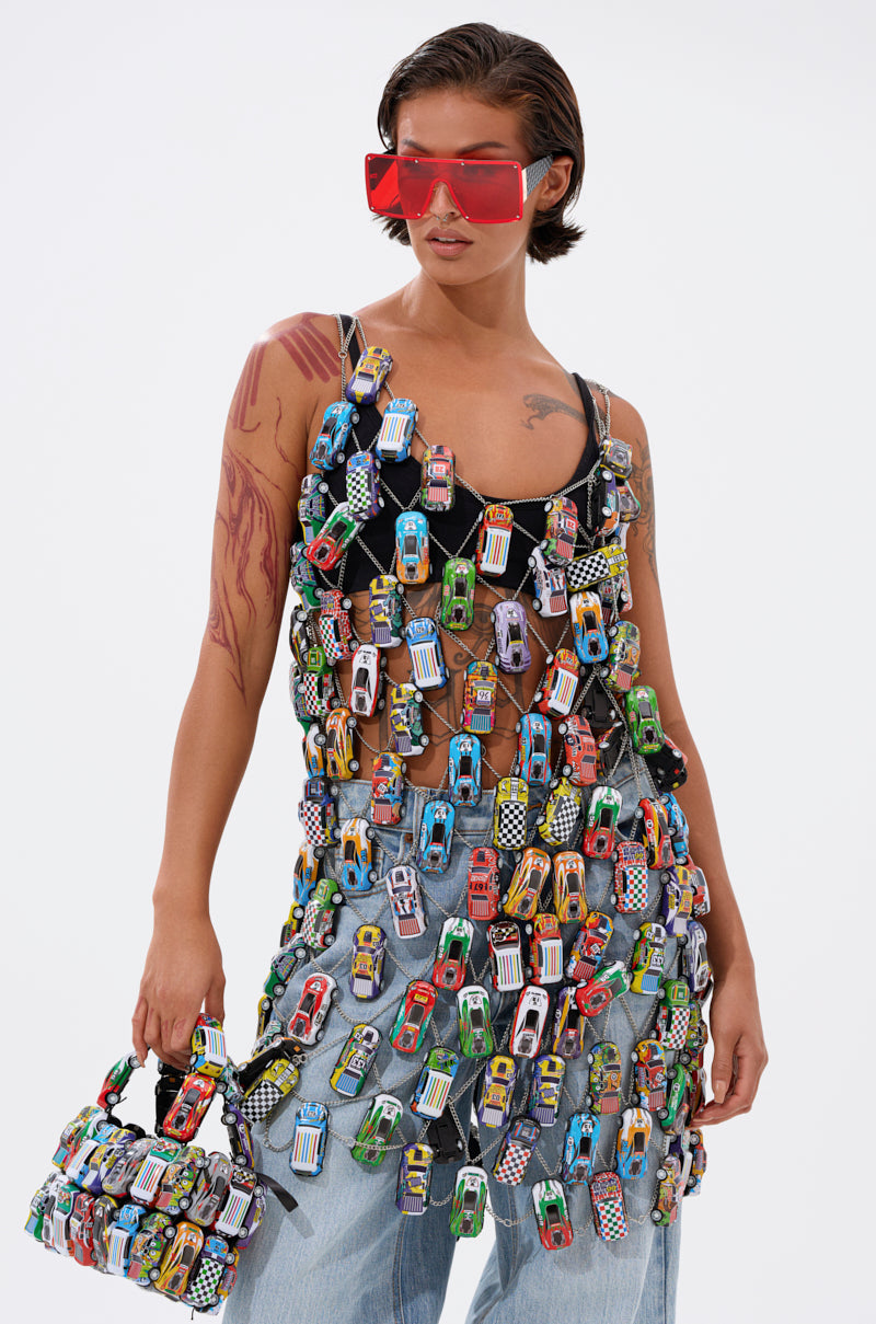 A person with short hair and red sunglasses wears the CAN'T CATCH ME CAR CHAIN MINI DRESS over blue jeans and a black top, carrying a handbag adorned with colorful toy cars, against a plain white background.