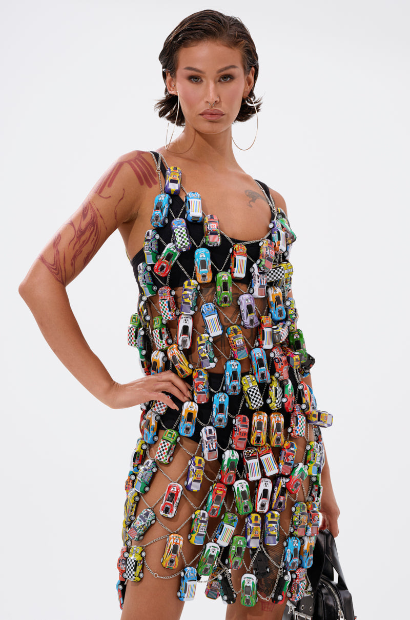 A woman poses confidently in the CAN'T CATCH ME CAR CHAIN MINI DRESS, featuring a sheer black fabric adorned with colorful toy cars, paired with slicked-back hair, hoop earrings, an arm tattoo, and a black handbag against a white background.