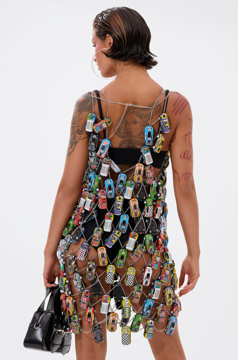 A tattooed person wears the CAN'T CATCH ME CAR CHAIN MINI DRESS over black undergarments, paired with a black handbag, wet-look hair, and large hoop earrings; they face away from the camera against a white background.
