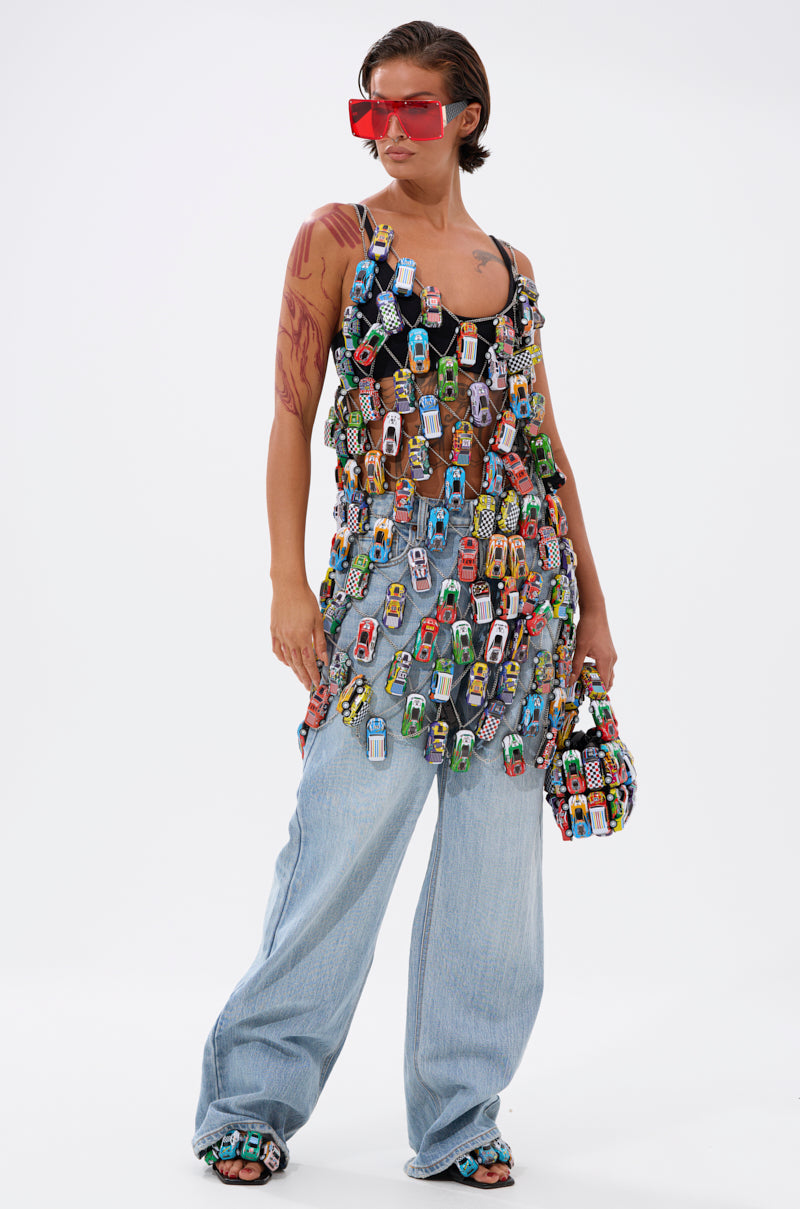A person poses confidently against a plain white background, wearing the CAN'T CATCH ME CAR CHAIN MINI DRESS styled with large red sunglasses, loose light blue jeans, and a matching toy car handbag.