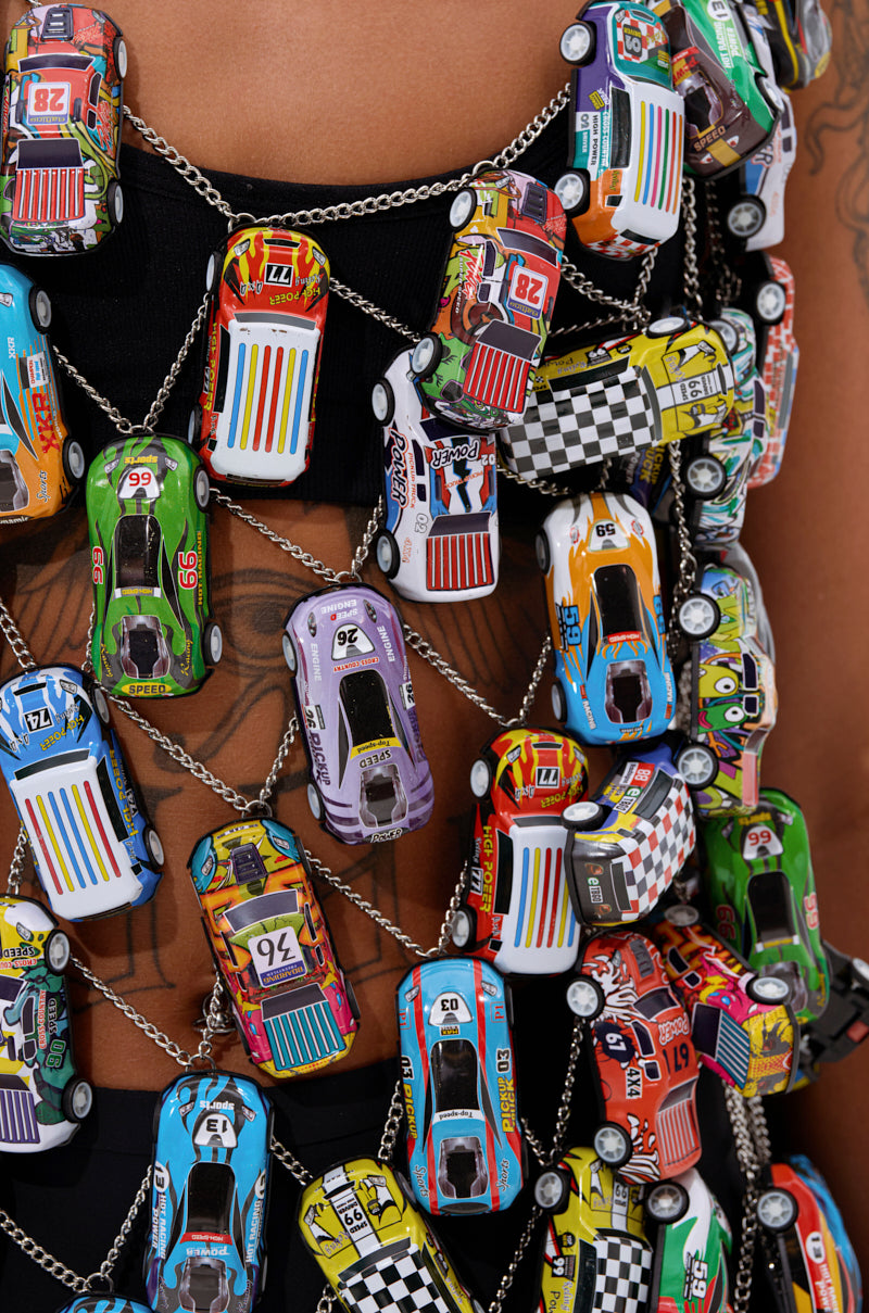Close-up of the CAN'T CATCH ME CAR CHAIN MINI DRESS, made from colorful toy race cars linked with metal chains and worn over a black top, revealing glimpses of tattooed skin through the dress's gaps.