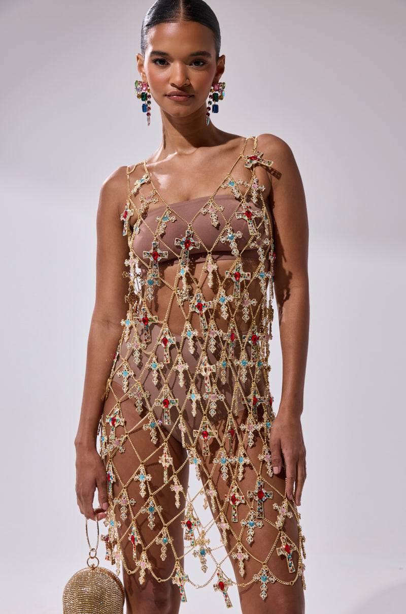 A woman wears the EVE CROSS CHAIN MINI DRESS, featuring a nude base with a gold mesh overlay adorned with colorful jewels. She accessorizes with a round gold bag, statement multicolored gem earrings, and her hair is styled in a sleek bun.