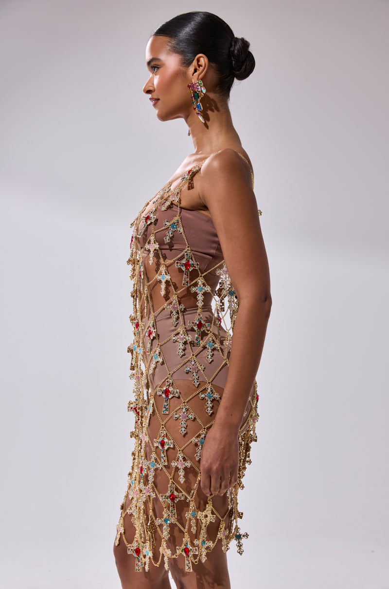 A woman in profile wears the EVE CROSS CHAIN MINI DRESS, a fitted nude mini dress embellished with colorful gems and cross-shaped ornaments. Her hair is styled in a sleek bun and she pairs the look with statement earrings.