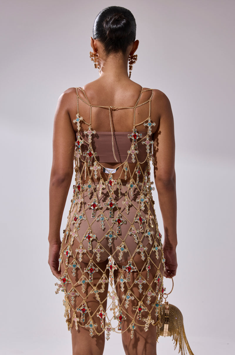 A person faces away in the EVE CROSS CHAIN MINI DRESS, featuring gold chains and colorful cross pendants over a nude slip. Their hair is in a sleek bun, and they carry a gold tassel handbag.