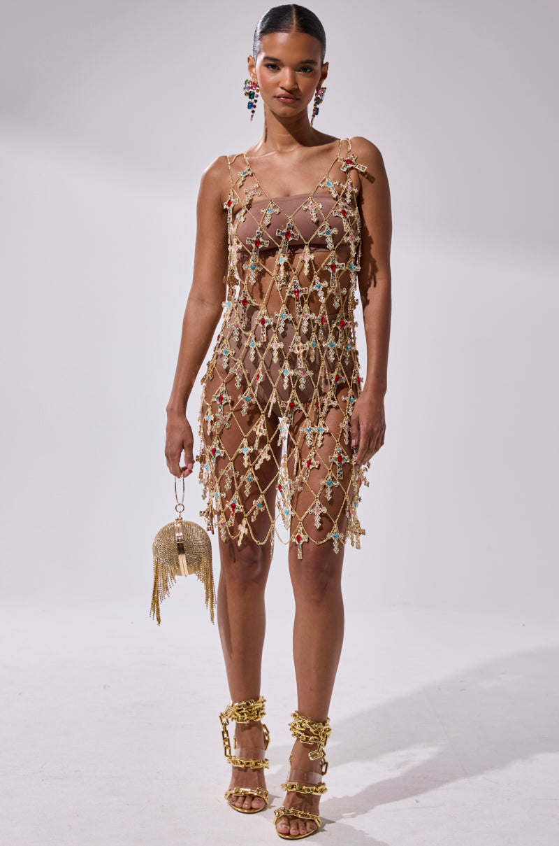 A woman poses against a white background in the EVE CROSS CHAIN MINI DRESS, a sleeveless mini adorned with gold chains and floral details over a nude slip. She accessorizes with gold chain sandals, statement earrings, and a gold fringed purse.