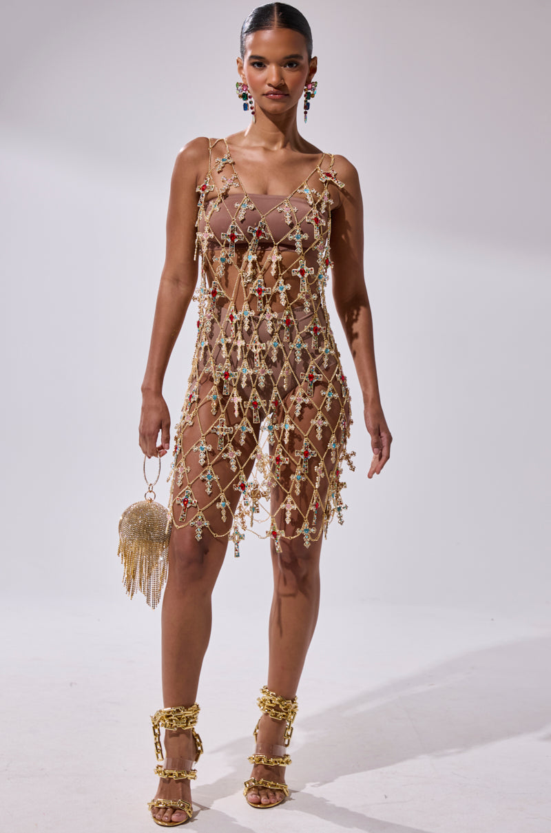 A woman stands against a plain background wearing the EVE CROSS CHAIN MINI DRESS with colorful beads over a nude slip, gold high-heeled sandals, statement earrings, and holding a gold fringe handbag.