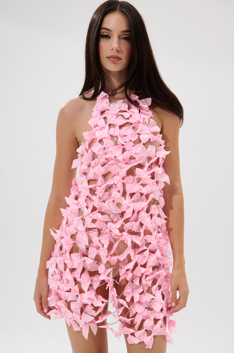 A woman with long, straight dark hair and a neutral expression poses against a plain background in the SLAP IT WITH A BOW CHAIN MINI DRESS—a short, sleeveless dress featuring pink bows for a semi-sheer, textured appearance.