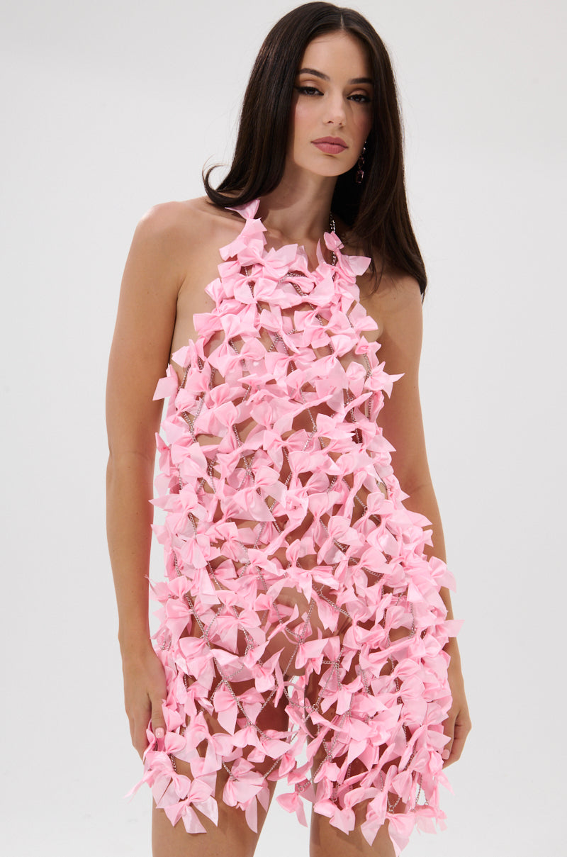 A woman with long dark hair models the SLAP IT WITH A BOW CHAIN MINI DRESS, featuring sheer fabric, pink satin bows, and an open-back design, while standing against a plain white background.