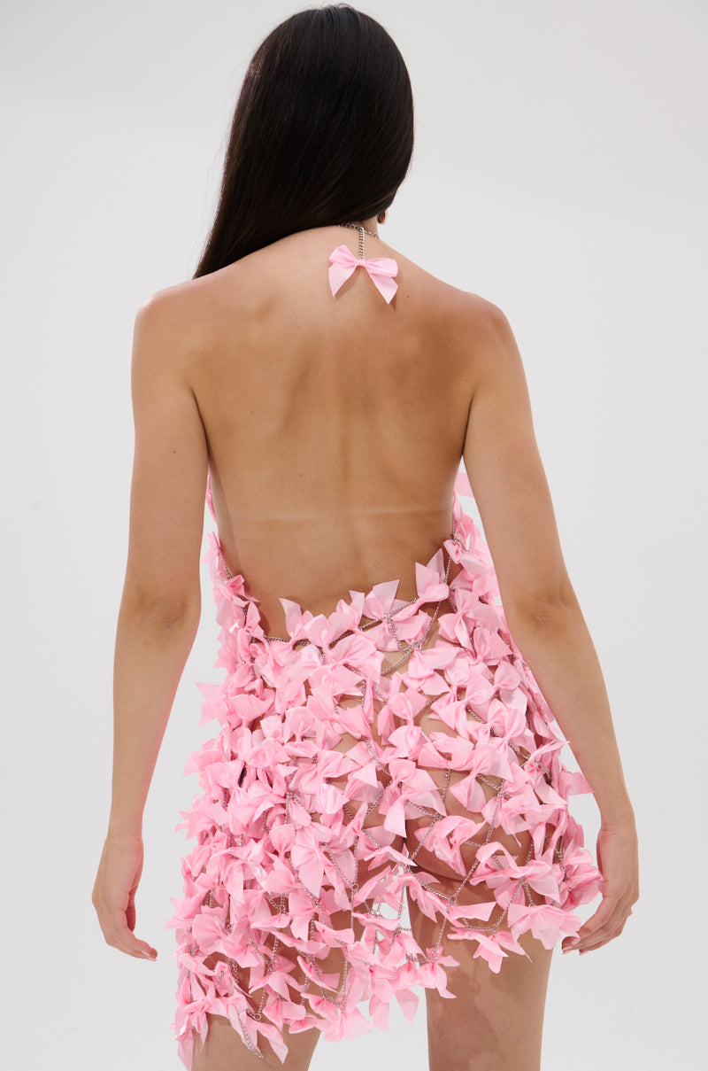A person with long dark hair is seen from behind, wearing the SLAP IT WITH A BOW CHAIN MINI DRESS—a sheer open-back dress featuring multiple pink satin bows and a matching neck bow. The plain background is light-colored.