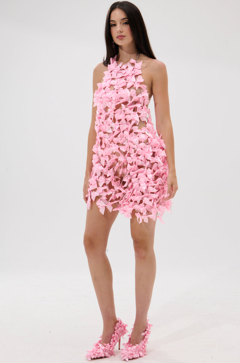 A woman poses against a white background wearing the SLAP IT WITH A BOW CHAIN MINI DRESS, featuring sheer fabric with pink floral petals and subtle pink satin bows. She pairs the look with matching petal heels, long straight hair, and a neutral expression.