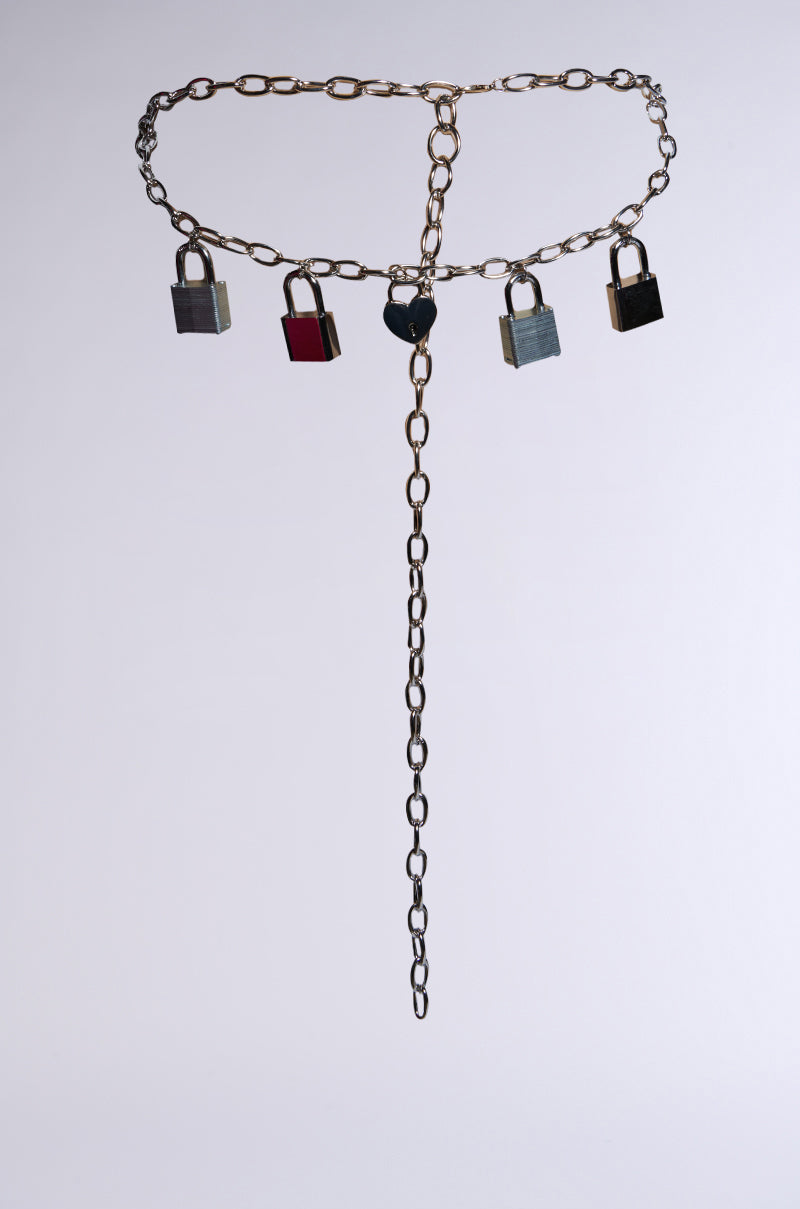 The HEART ON LOCK CHAIN BELT features a bold, metal chain link design shaped as a belt, hanging vertically with an extension and secured by five silver padlock charms. The background is plain and light-colored. 1