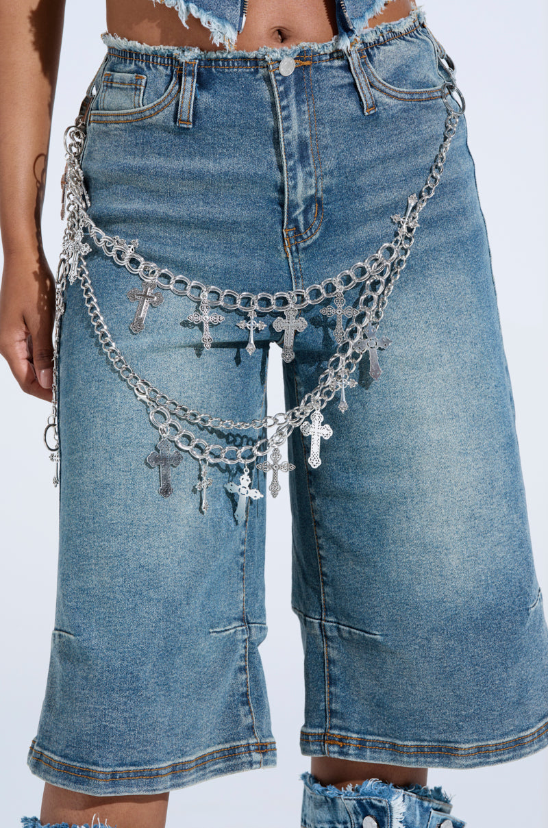 Distressed denim shorts with raw edges pair perfectly with the bold ULTIMATE CROSS CHAIN BELT featuring silver chains and crosses, creating a festival-ready look captured from waist to just below the knee.