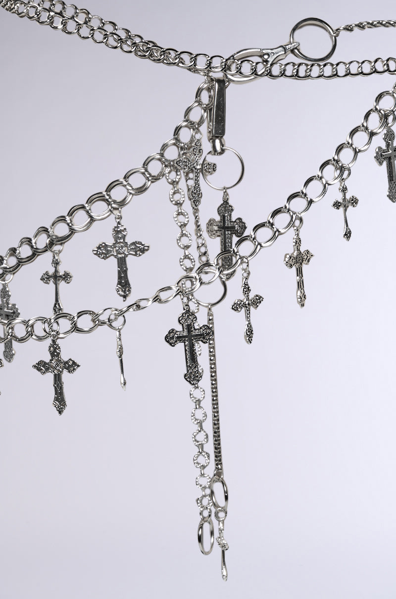 The ULTIMATE CROSS CHAIN BELT features silver cross chains with various sized dangling crosses on a gray backdrop. Its intricate design includes multiple loops and edgy hardware clips, delivering a festival-ready look with decorative flair.
