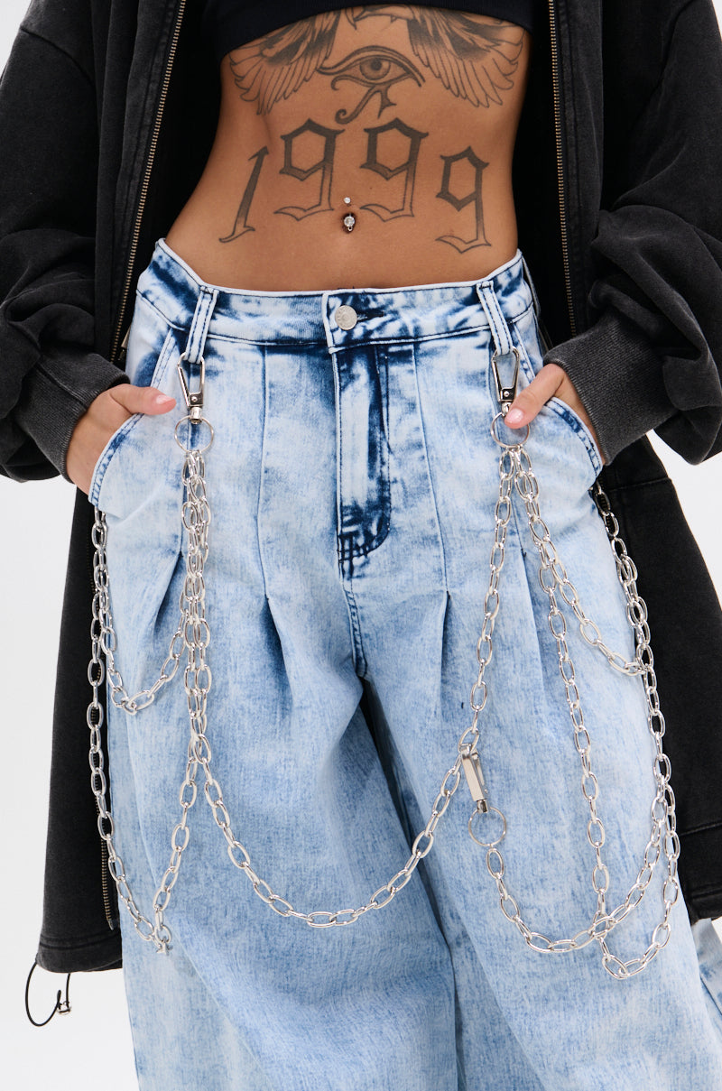 A trendsetter in light blue acid-wash jeans sports the PROUD OF YOU CLIP-ON PANT CHAIN, paired with a dark jacket and showing a "1999" plus eye tattoo on their stomach. 0