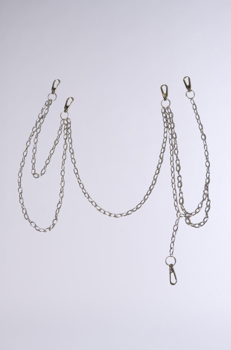 The PROUD OF YOU CLIP-ON PANT CHAIN, in silver with four statement clasps, elegantly drapes against a light gray background. It features three overlapping loops for a harness-like cascading effect. 1