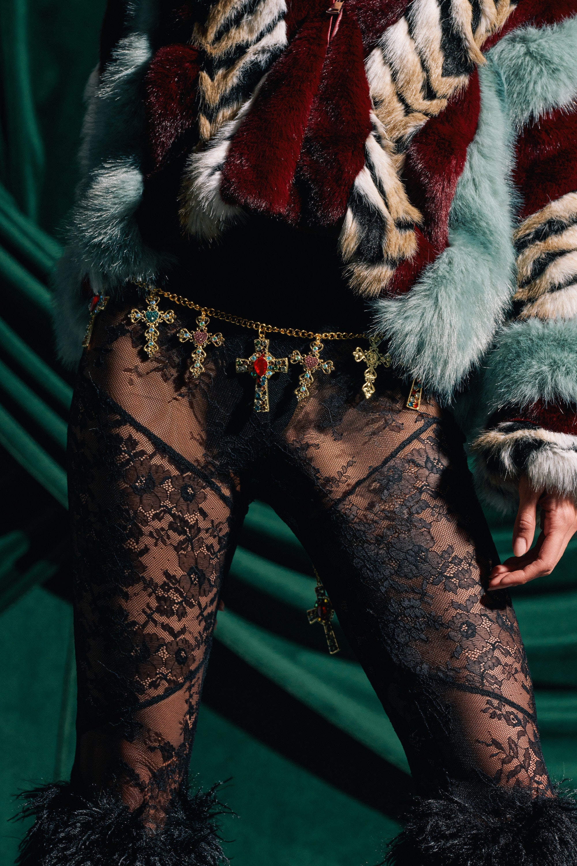 Wearing a faux fur animal print jacket, black lace pants, and the IN MY FEELINGS CHAIN BELT with cross pendants, a person stands before green draped fabric. 0