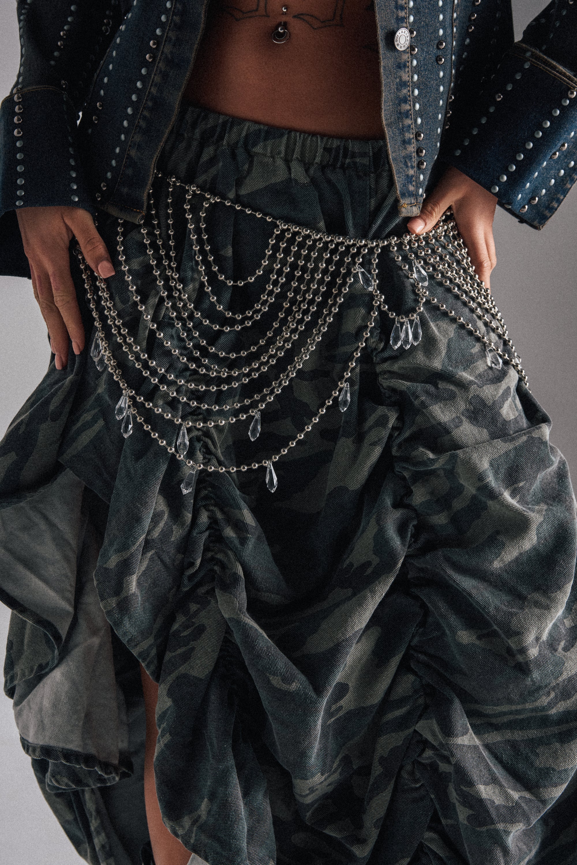 A person wearing a dark jean jacket with silver studs and a camo-patterned skirt accessorized with the DRIPPIN CHAIN BELT draped in silver-tone ball chains over the hips, hands resting on their waist. 0