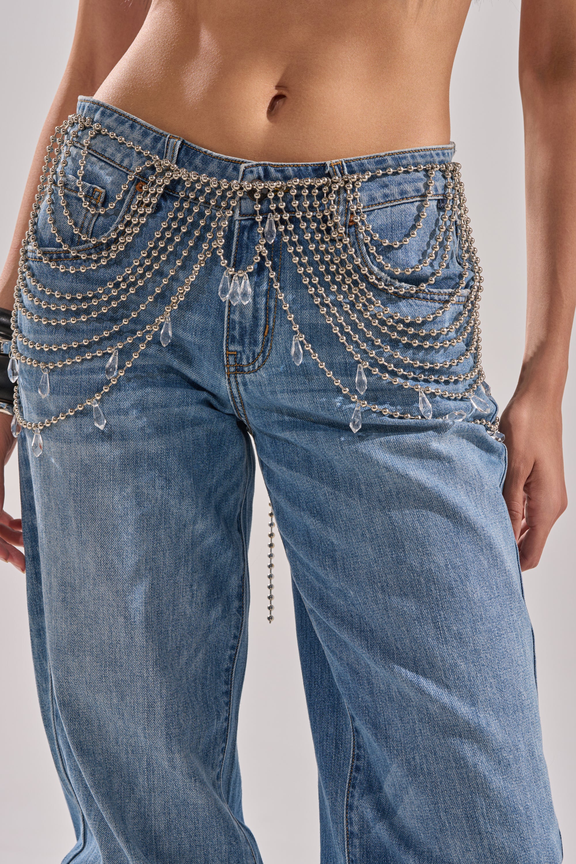 A person wearing loose blue jeans styled with the DRIPPIN CHAIN BELT, a silver-tone layered accessory draped across the hips and adorned with dangling crystal charms. The person's midriff is visible. 1