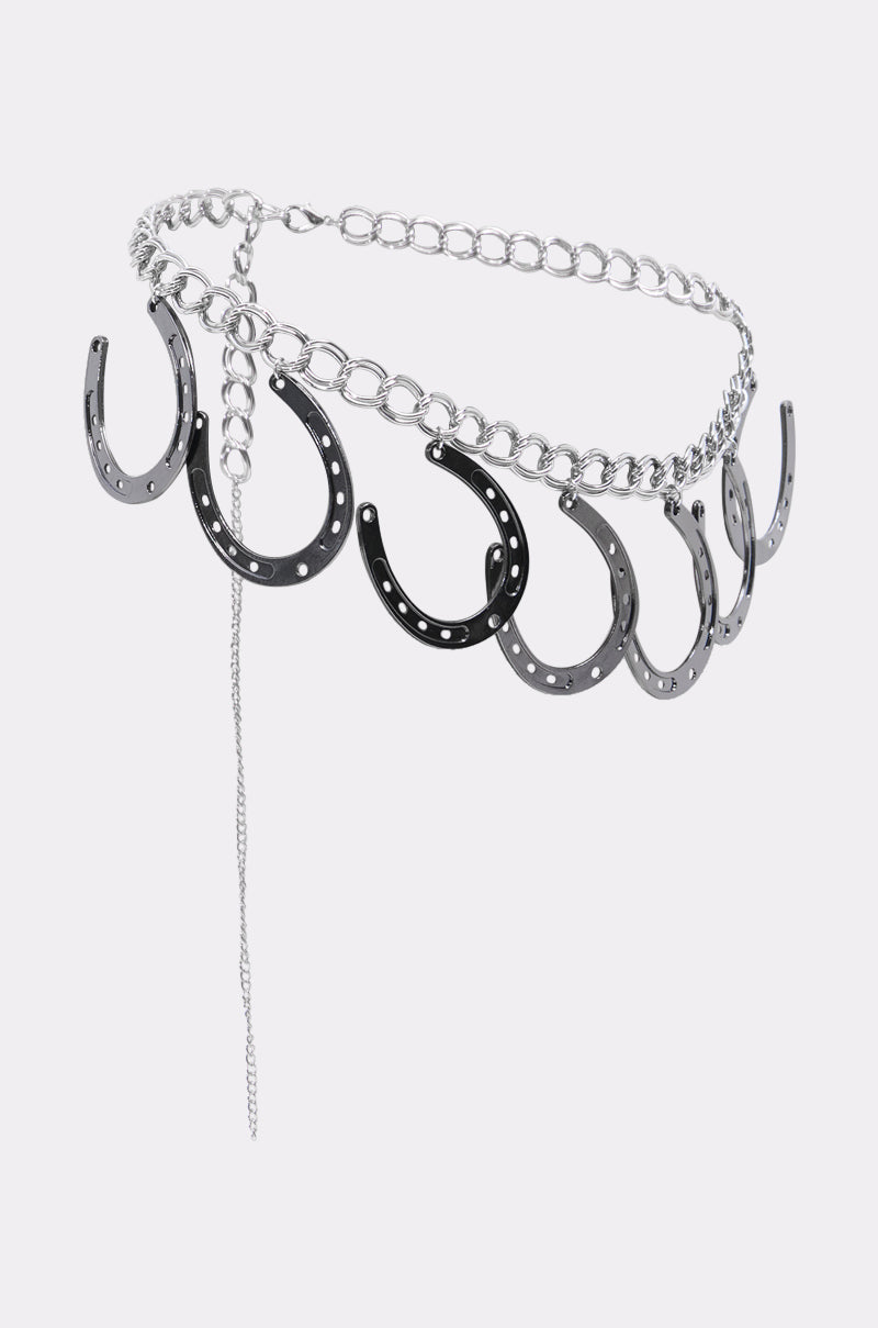 The TEST MY LUCK HORSESHOE BELT is a silver Western-inspired accessory featuring black and silver horseshoe pendants, ideal for rodeo looks, shown against a light gray background. 4