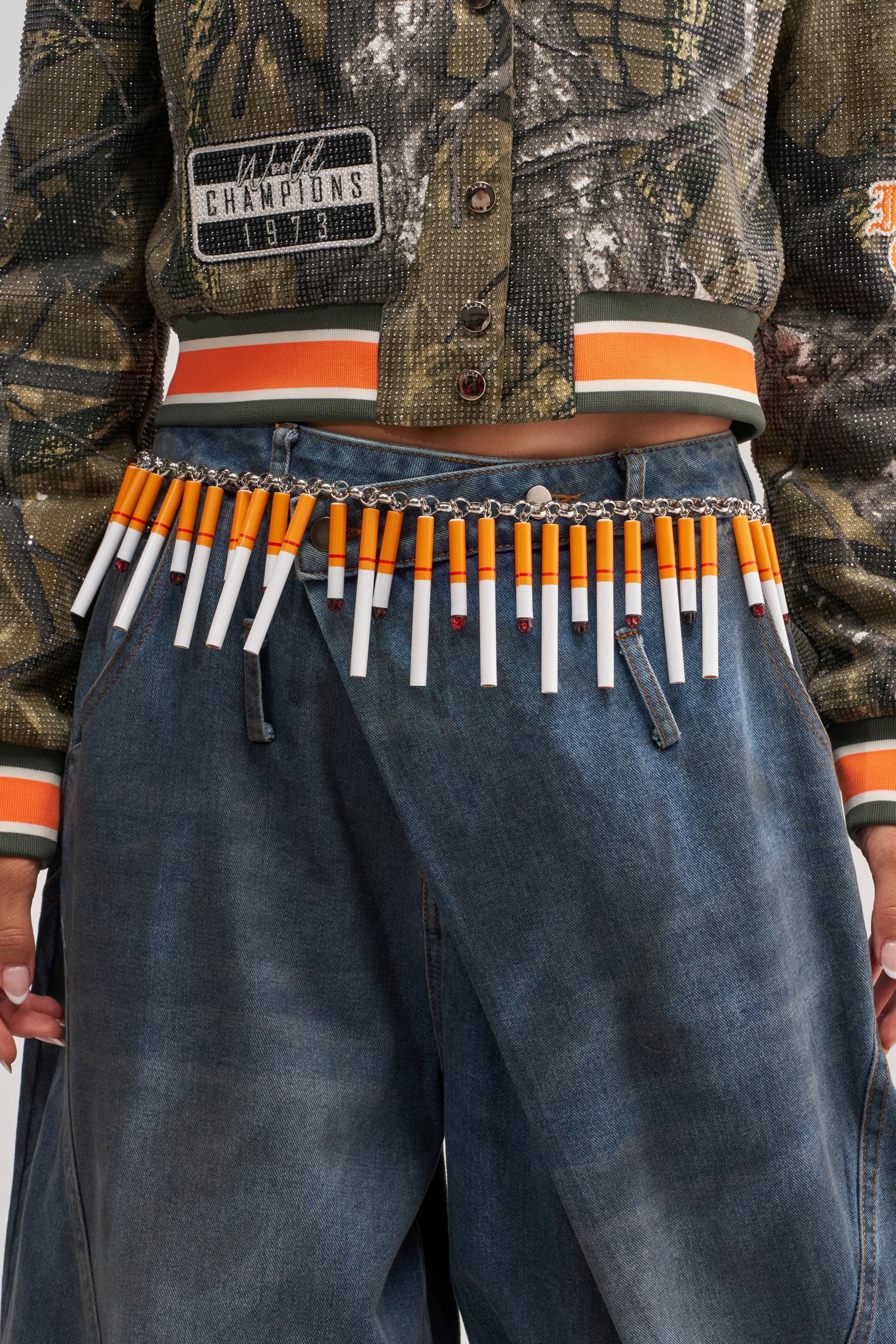 A person wearing blue jeans, a camouflage jacket with patches, and the CAN I LIGHT UP BELT—a striking festival accessory crafted from cigarettes strung together and inspired by bold cigarette embellishments.
