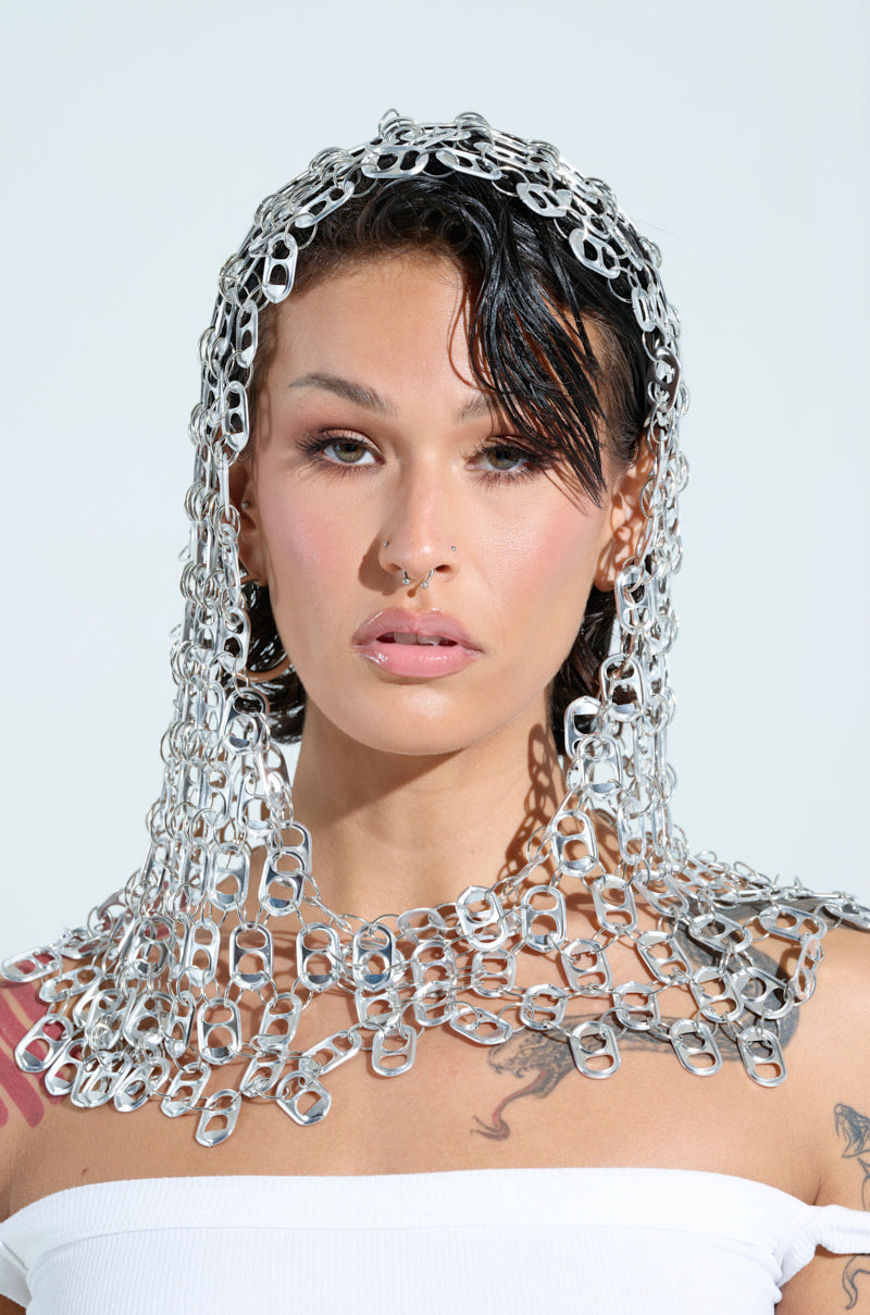 A person in a white top sports a WAYNE POP TAB HOOD headpiece and shoulder covering crafted from metal tabs, creating an avant-garde vibe. They have a septum piercing and shoulder tattoos, while the plain background accentuates their unique jewelry.