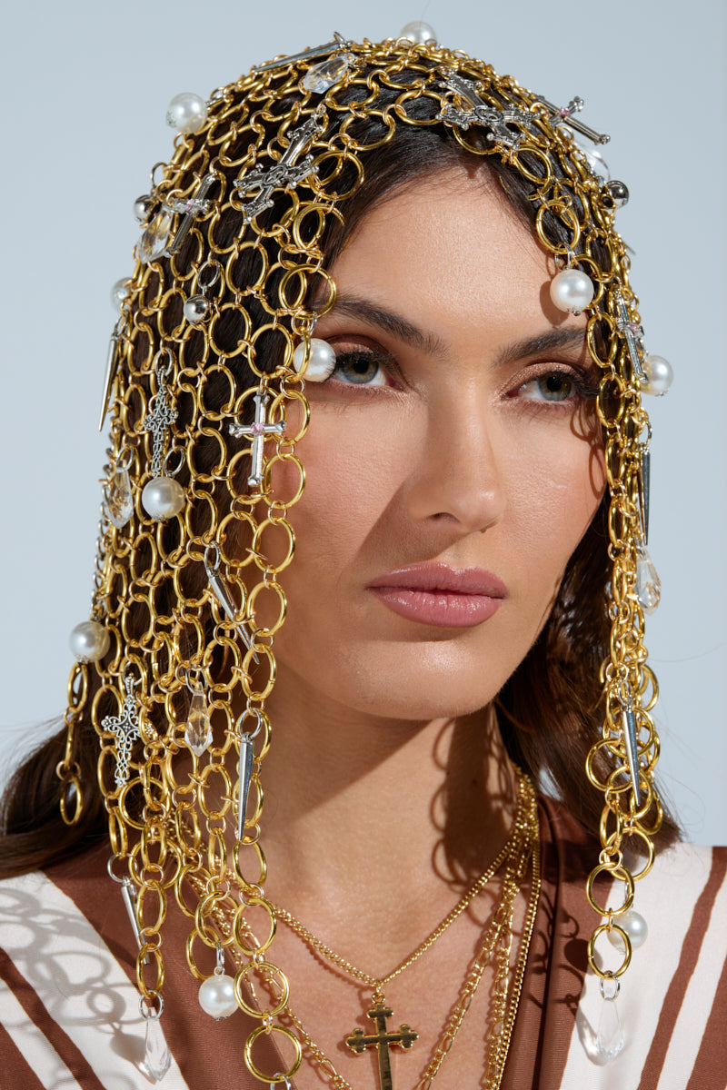 A woman with long dark hair wears the ALWAYS BLESSED HEADPIECE adorned with gold chains, pearl embellishments, and cross pendants. Silver chain link necklaces gracefully layer her neck, enhancing her neutral expression against a light background. 0