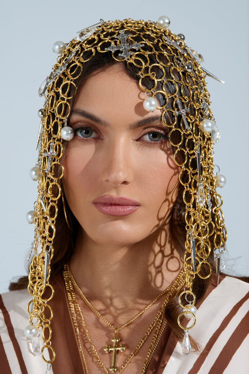 A person with long hair wears the ALWAYS BLESSED HEADPIECE, featuring pearl embellishments and gold chains that elegantly drape over their head. With a neutral expression, they layer gold necklaces over white and brown attire against a light background. 2
