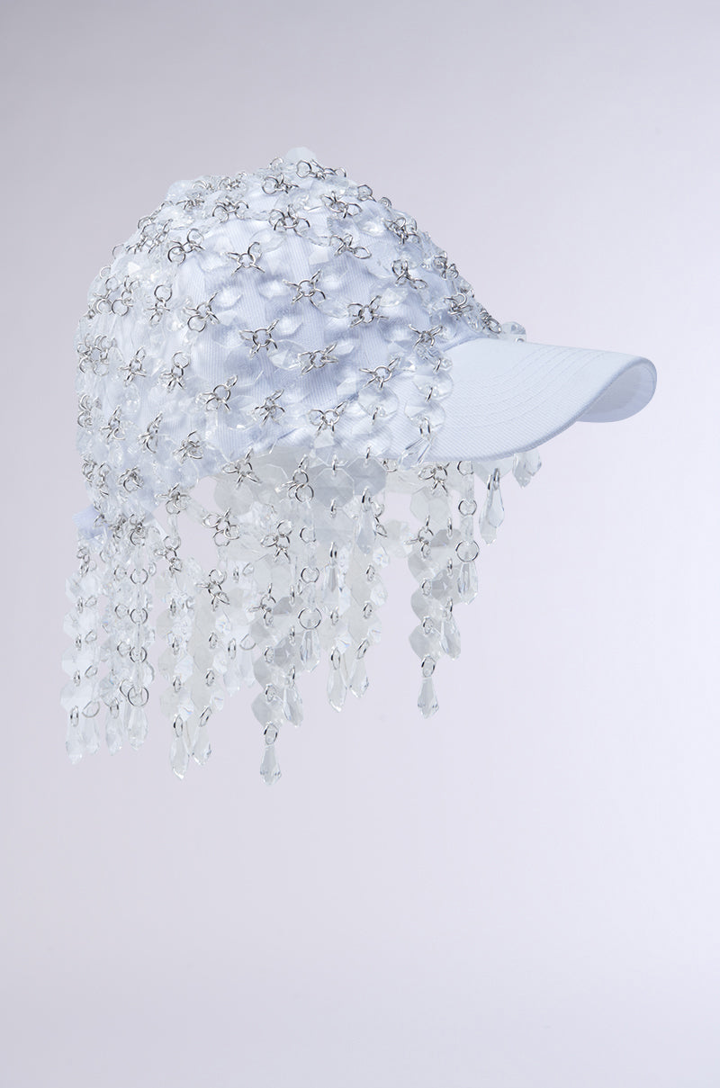 The IN THE CLEAR BEADED HAT is a white baseball cap with a veil-like net of clear beads and crystals draping over the front and sides, shown against a plain light gray background.