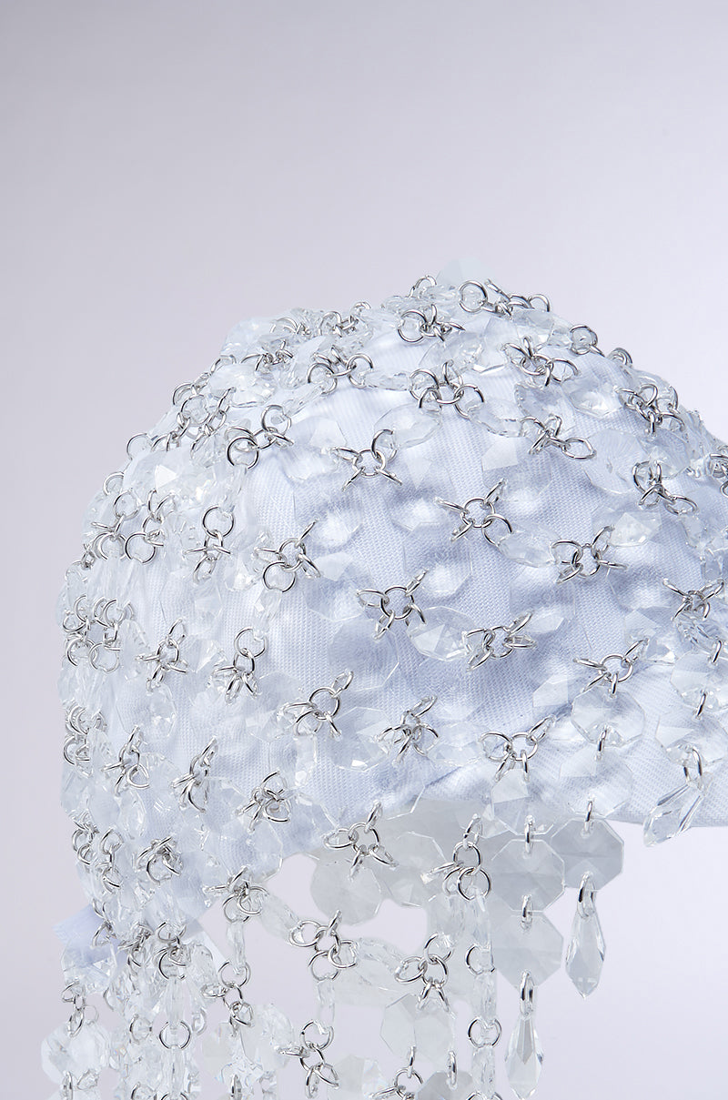 A close-up of the IN THE CLEAR BEADED HAT showcases its white textured surface, metallic hardware, and clear crystal-like beads, creating a shimmering, layered effect against a pale background.
