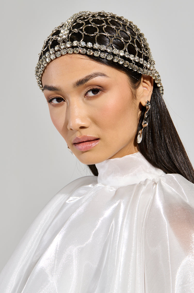 Wearing a TOO LUXE RHINESTONE CAP and a white satin garment, the woman gazes toward the camera. Her long, dark hair is sleek, with metallic rhinestone statement earrings adding sparkle. The plain background enhances her elegant attire and accessories. 0