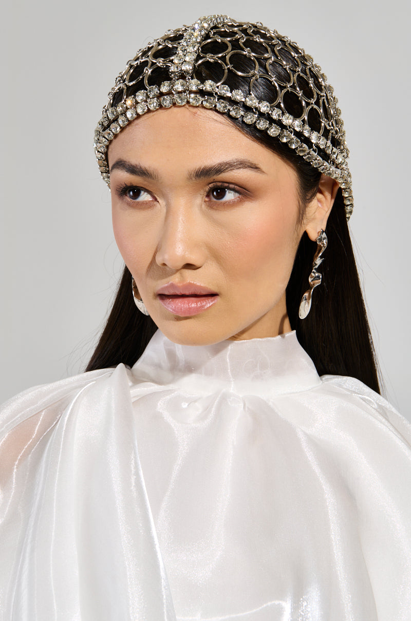 A woman wearing the TOO LUXE RHINESTONE CAP, adorned with circular patterns and gemstones that shimmer like metallic rhinestones. She has a white, high-neck blouse, large dangling earrings, and displays a serious expression while looking slightly to her left against a neutral background. 1