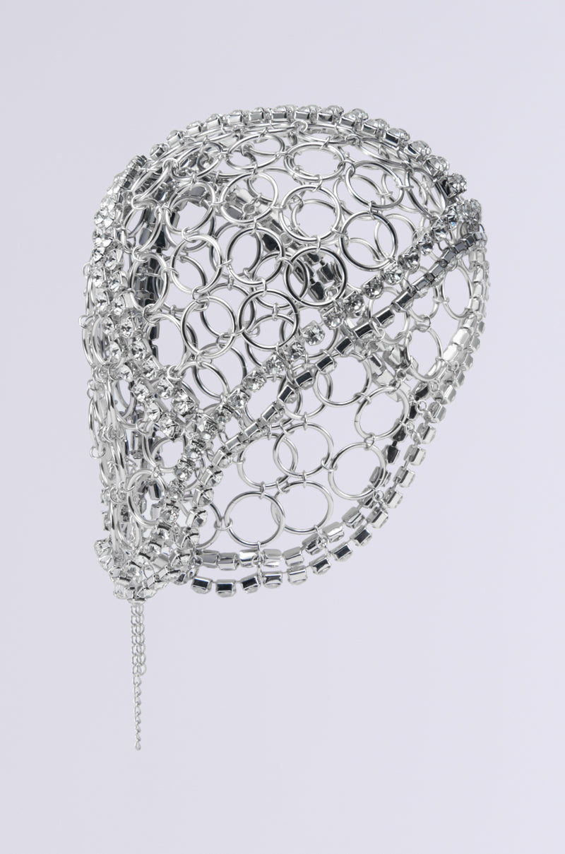 The TOO LUXE RHINESTONE CAP is a silver metallic headpiece featuring interconnected circular rings and small chains that create an intricate, open-patterned design. The headpiece showcases a structured, abstract shape set against a plain white background. 3