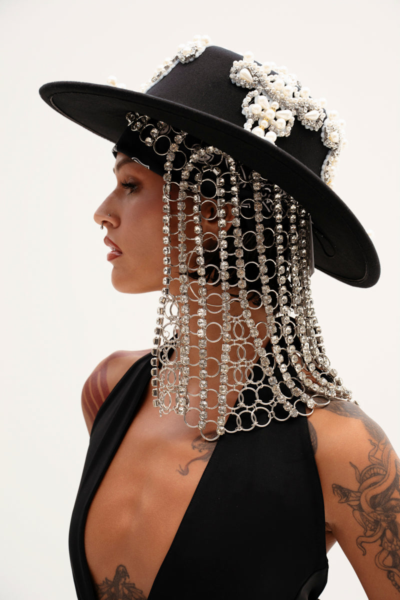 Embodying queen sh*t vibes, a person dons the QUEEN SH*T BLING HEADPIECE—a black wide-brimmed hat adorned with silver rhinestones and floral decorations. In all black, tattoos peek from their arm as they gaze sideways against a light backdrop, exuding striking elegance.