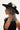 Embodying queen sh*t vibes, a person dons the QUEEN SH*T BLING HEADPIECE—a black wide-brimmed hat adorned with silver rhinestones and floral decorations. In all black, tattoos peek from their arm as they gaze sideways against a light backdrop, exuding striking elegance.