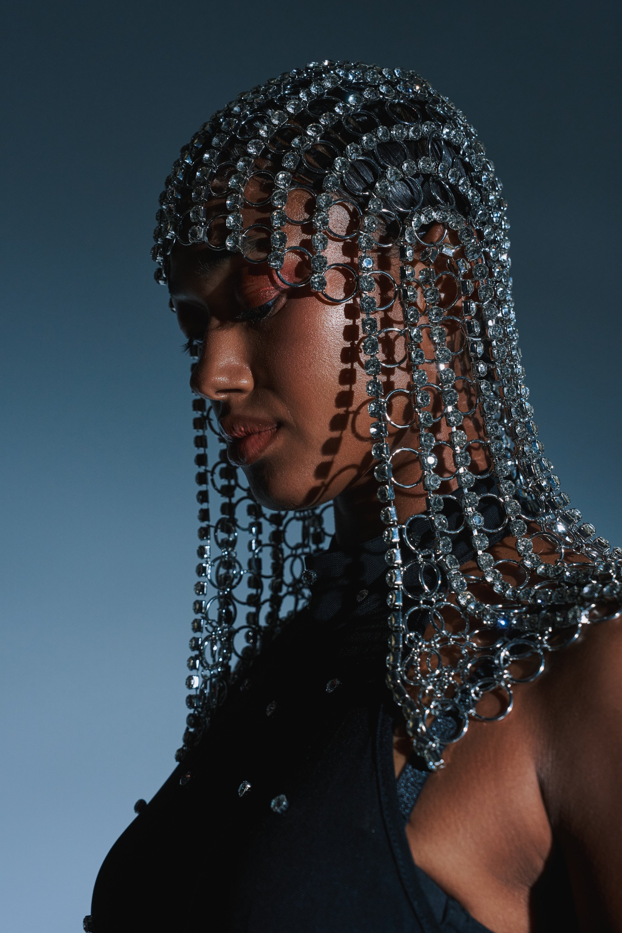 A woman in profile wears the QUEEN SH*T BLING HEADPIECE—silver chains and beads catching dramatic light on her smooth skin and dark sleeveless top—set against a blue-gray background.