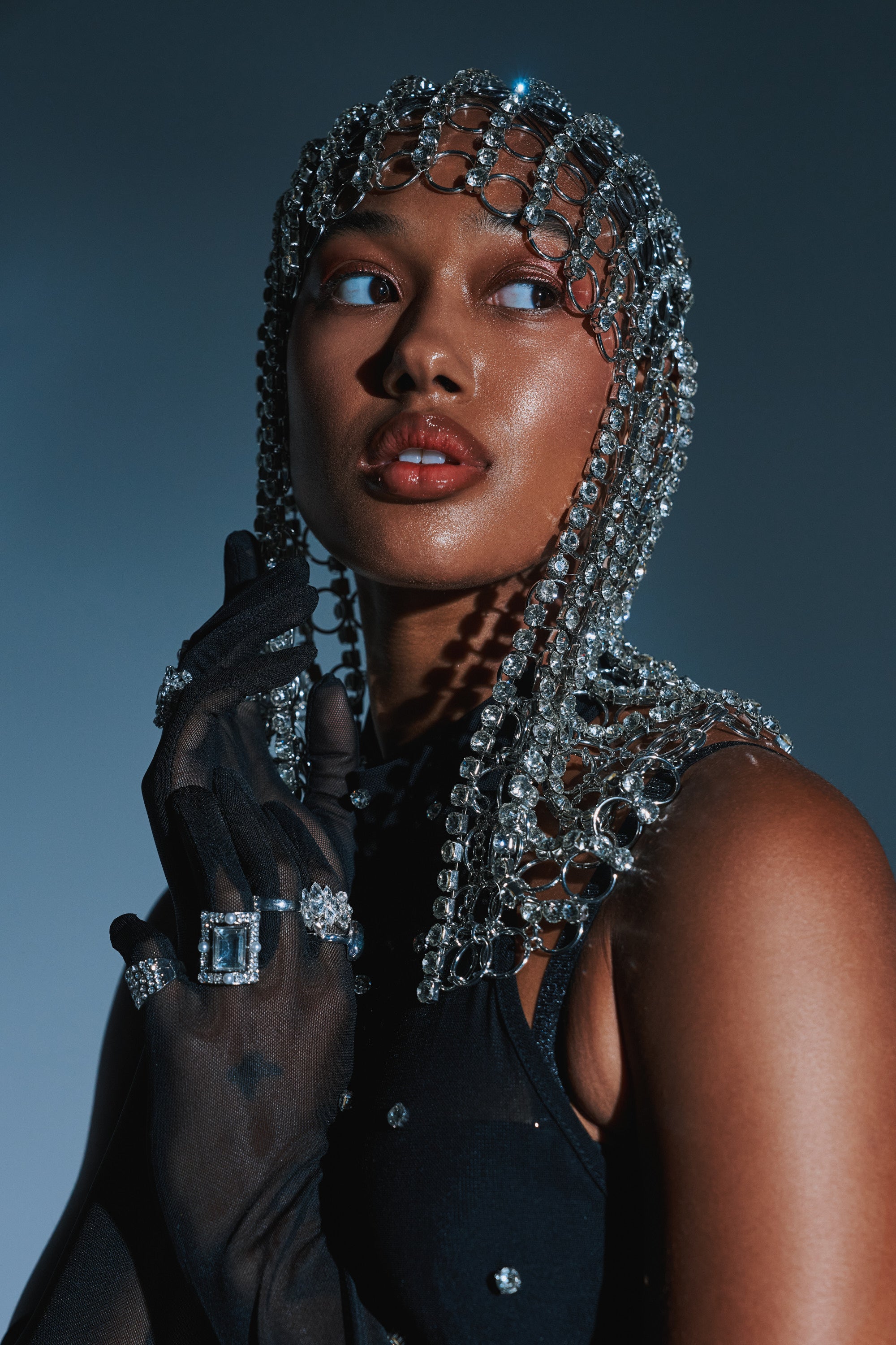 A woman poses against a dark backdrop, wearing the QUEEN SH*T BLING HEADPIECE adorned with rhinestone chains and crystals, sheer black gloves, sparkling rings, and gazing to the side with parted lips and a dramatic expression.