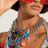 A person flaunts oversized red sunglasses and the ROLL THE DICE CHARM CHOKER, adorned with colorful bear charms hanging from multiple chains. The silver metallic base shimmers in the light, and an adjustable lobster clasp closure guarantees a perfect fit. They also showcase a nose ring and visible tattoos against a simple, light background.