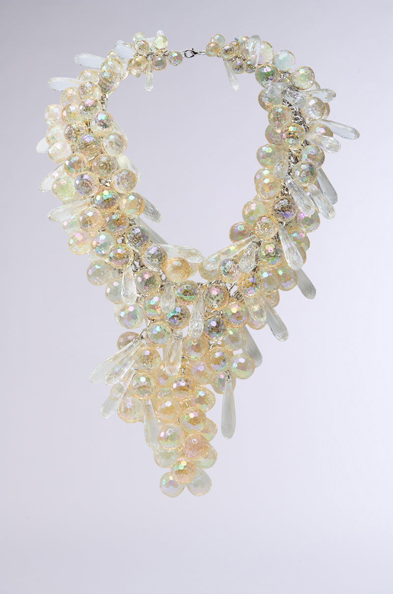 Meet the ICONIC BEAUTY NECKLACE: a statement piece featuring clusters of iridescent beads and teardrop crystals cascading beautifully. Shimmering against a gray backdrop, it's complemented by a layered silver chain for added allure.
